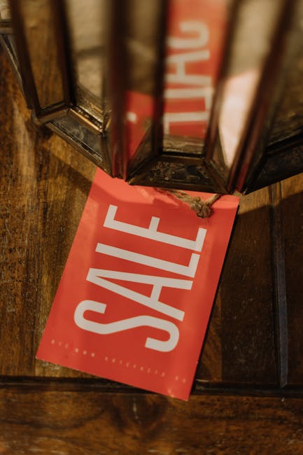 A red rectangular sale sign with bold white capital letters spelling 'SALE' is positioned on a wooden floor beneath a glass display case. The sign is slightly crumpled around the edges and partly tuck
